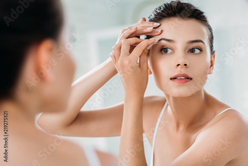 beautiful girl correcting eyebrows with tweezers and looking at mirror in bathroom photo
