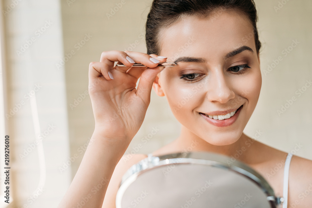 smiling young woman correcting eyebrows with tweezers and looking at mirror smiling young woman correcting eyebrows with tweezers and looking at mirror