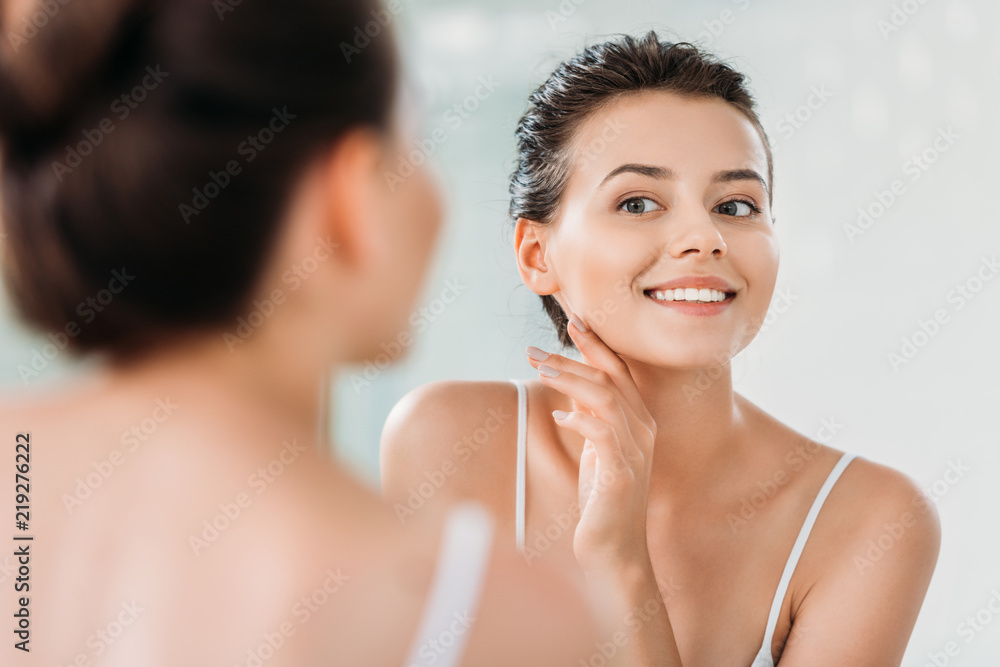 beautiful smiling young woman touching skin and looking at mirror in bathroom - obrazy, fototapety, plakaty 
