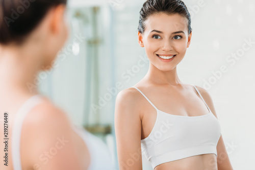 selective focus of beautiful smiling girl looking at mirror in bathroom