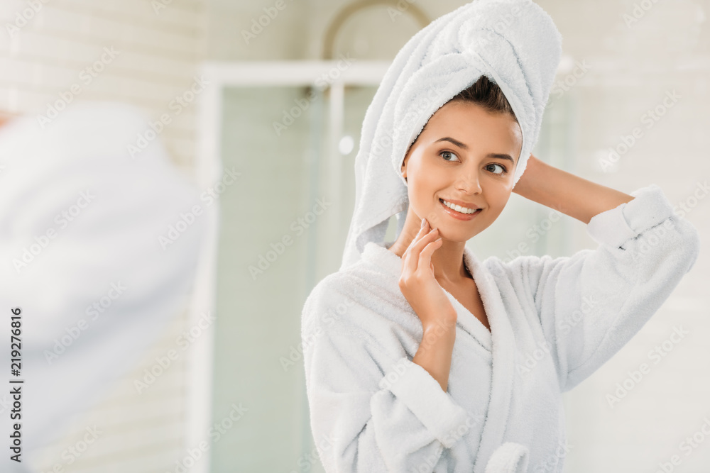 beautiful smiling young woman in bathrobe and towel on head looking at mirror in bathroom