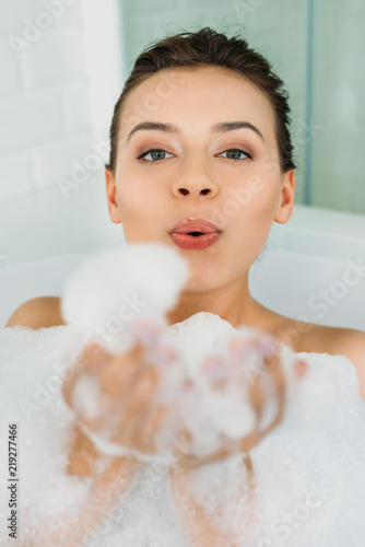 beautiful young woman blowing foam and looking at camera in bathroom