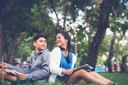 Happy Asian school boy and girl turning heads to each other while sitting on green grass in schoolyard and reading books