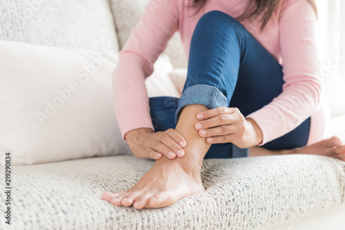 Closeup woman sitting on sofa holds her ankle injury, feeling pain. Health care and medical concept. photo
