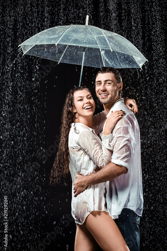 side view of happy couple in white shirts standing under umbrella under raindrops isolated on black