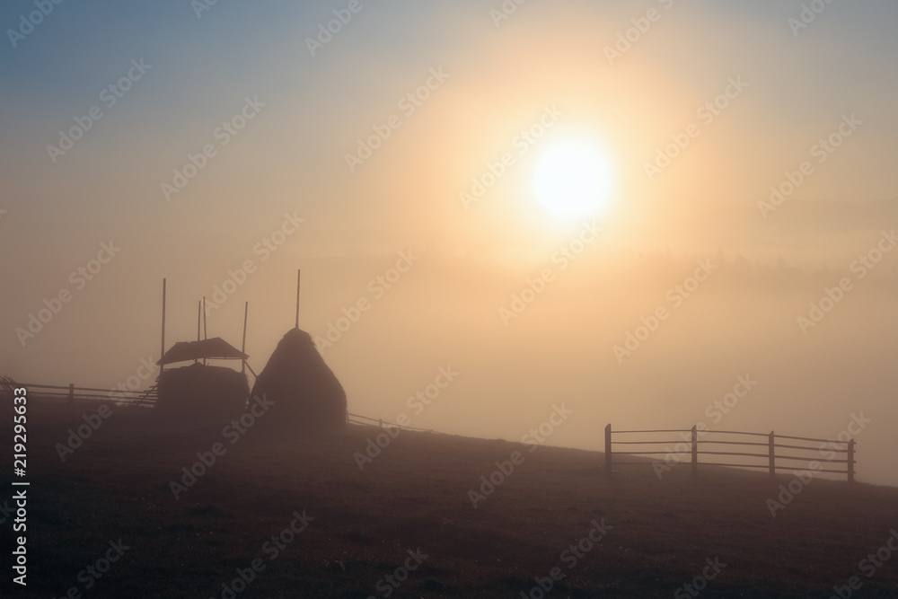 The bright disk of the sun illuminates with a warm orange light the mountain meadow where loose stacked hays built around a central pole stand, which are shrouded in a light mist.