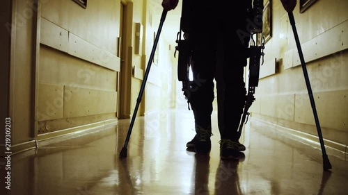 Close up silhouette of the legs of disable man in the robotic exoskeleton walking through the corridor with support of a doctor. Then camera moving to the face and back to the legs