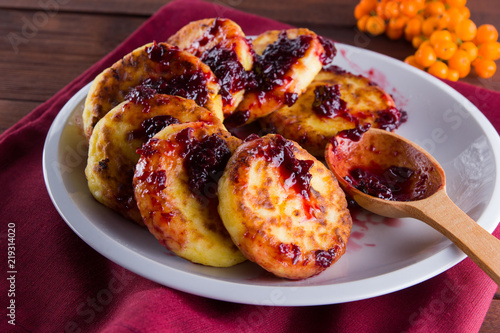 Cottage cheese pancakes on a wooden background. Traditional Ukrainian dish - syrniki with jam on a red napkin. Pancakes with cottage cheese on a white plate with honey and rowan berries