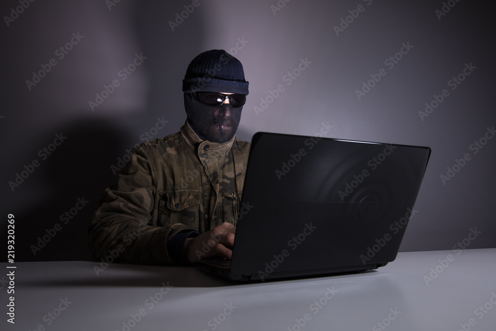 Network criminal man examining a laptop Computer. The man has a camouflage jacket, sunglasses, and balaclava. The photo is underexposed. Network criminal man examining a laptop Computer. The man has a camouflage jacket, sunglasses, and balaclava. The photo is underexposed.