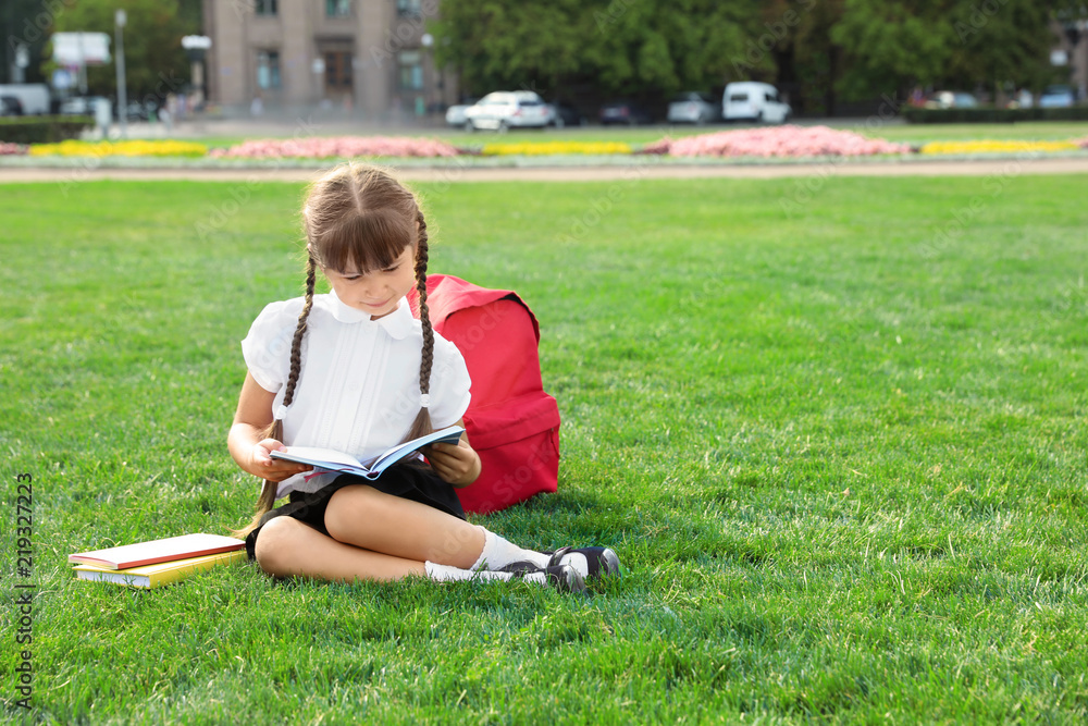 Cute girl with school stationery reading book on green lawn outdoors Cute girl with school stationery reading book on green lawn outdoors