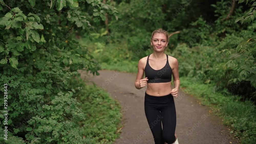 Attractive slim young woman jogging in a summer park in black sports bra. Front view Left to right pan slow motion medium shot Attractive slim young woman jogging in a summer park in black sports bra. Front view Left to right pan slow motion medium shot