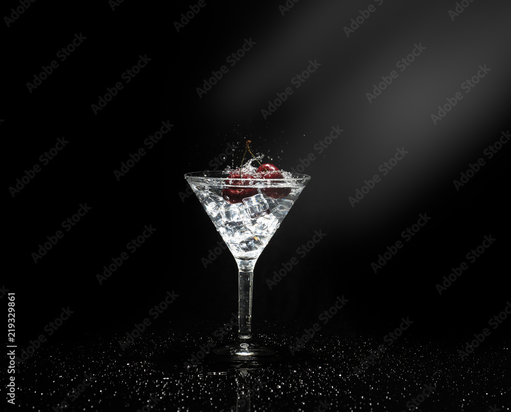 Close up view of splash water with falling cherry in a martini glass among ice in black background with flare.