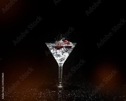 Close up view of splash water with falling cherry in a martini glass among ice in black background foreground.