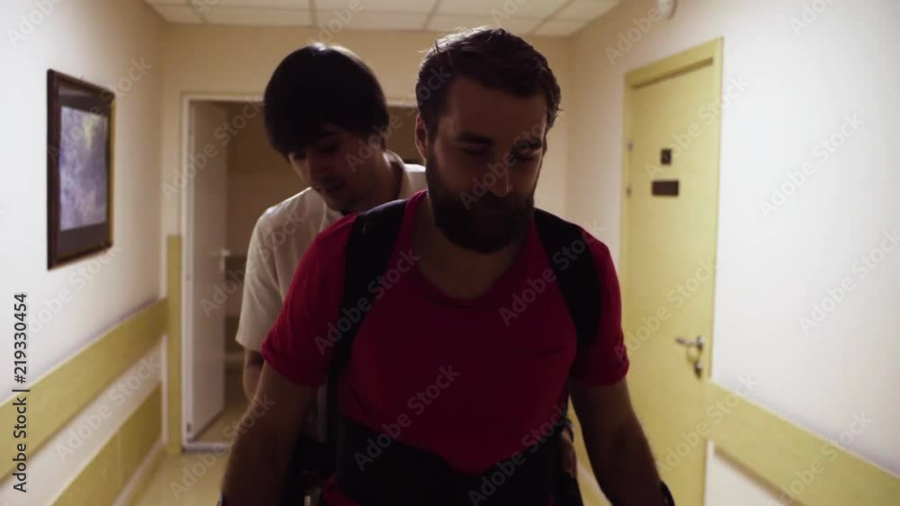Portrait of young disable man walking in the robotic exoskeleton through the corridor of the rehabilitation clinic. Doctor walking behind him. Portrait of young disable man walking in the robotic exoskeleton through the corridor of the rehabilitation clinic. Doctor walking behind him.
