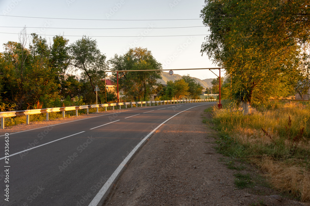 asphalt road, striped black and white road barrier in the village during sunset
