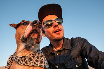 Spoiled self portrait concept: man trying to make a selfie with his funny dog. Young male person and staffordshire terrier posing for a selfie outdoors
