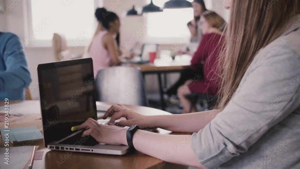 Close-up of young female freelance worker with smart watch typing on laptop by the table in busy modern coworking space. Close-up of young female freelance worker with smart watch typing on laptop by the table in busy modern coworking space.