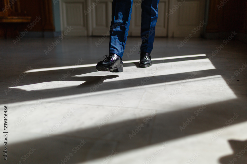 Legs of a man in an expensive suit and black shoes during a step. A person walks along the corridor of the living room or hotel marble floor under the sunlight. Legs close up. Business concept