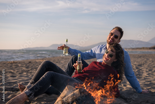 Young Couple Sitting On The Beach beside Campfire drinking beer