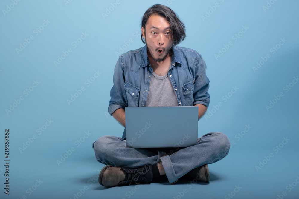 Young bearded man sit down on floor using computer in blue background.