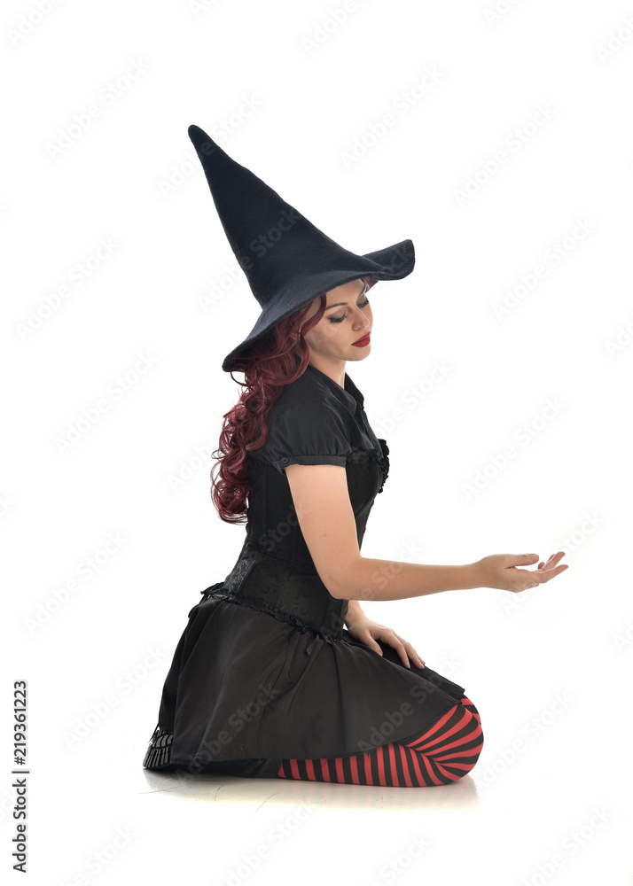 Fototapeta premium full length portrait of red haired girl wearing black witch costume and pointy hat. seated pose, isolated on white studio background.