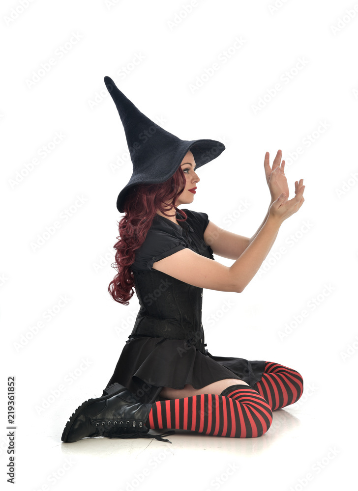 Naklejka premium full length portrait of red haired girl wearing black witch costume and pointy hat. seated pose, isolated on white studio background.