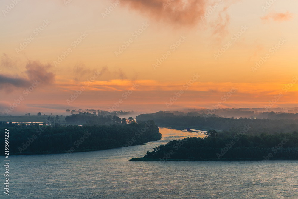 Fototapeta premium River flows along shore with forest under fog. Channel of river flows around island. Orange glow in dawn sky reflected on water. Colorful morning mystical atmospheric landscape of majestic nature.
