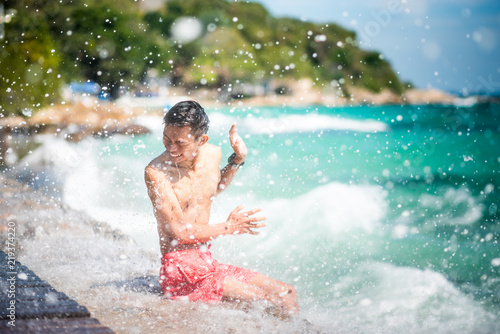 Smiling young Asian man sits on a sea pier with the excited face & expressive hand gesture being wet under shower of splash after a large wave hit. Scene of happiness at sunny day. Koh Samet, Thailand