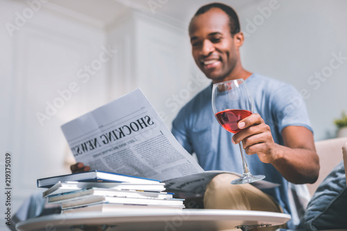 Time spending. Cheerful smiling man reading articles while drinking red wine