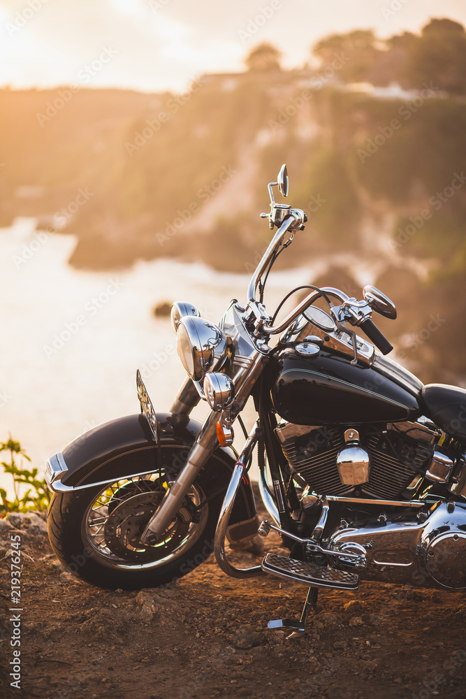 Fototapeta premium Stary rocznika motocyklu pozycja na krawędzi falezy w ciepłym świetle słonecznym przy wschodem słońca, błyszczący szczegóły roweru zakończenie