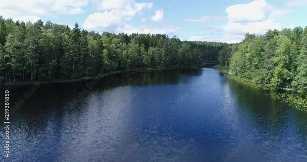 Drone flying forward and rising over calm lake near shoreline over tree line during sunny summer day. White clouds and blue sky reflecting from the water. Haukkalampi. Nuuksio. Espoo. Finland Drone flying forward and rising over calm lake near shoreline over tree line during sunny summer day. White clouds and blue sky reflecting from the water. Haukkalampi. Nuuksio. Espoo. Finland