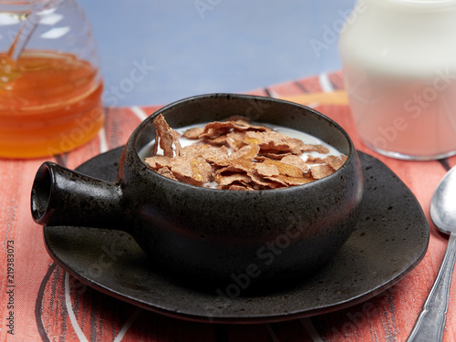 A bowl of oat bran flakes with milk and honey set on a red napkin, on a blue tablecloth, with a jar of honet and a jug of milk photo