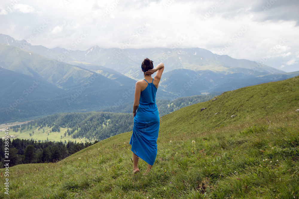 woman in a blue long dress stands on top of a mountain in the summer on a green meadow
