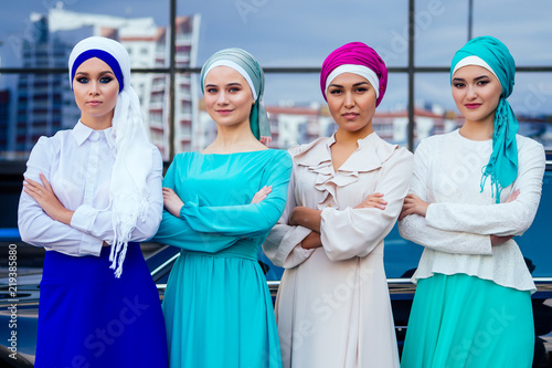 a group of four beautiful perfect makeup business women in muslim dress and turban on head standing in front of the car skyscraper street background