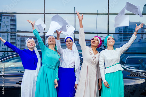 group of five strong beautiful and young Muslim business women fashion look stylish long dress and the turban on head holding paper ,car and skyscraper street background.good deal success teamwork