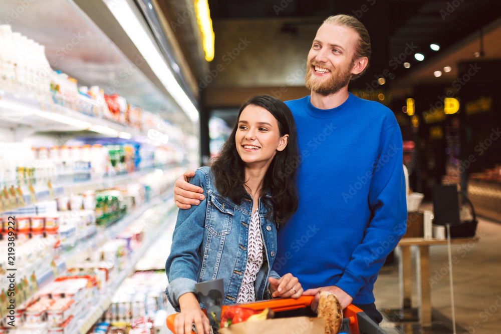 Young cheerful couple with trolley full of products happily choosing dairy in supermarket