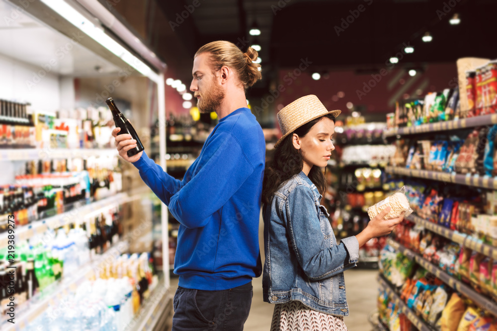 Young beautiful couple standing back to back thoughtfully choosing products while spending time in modern supermarket