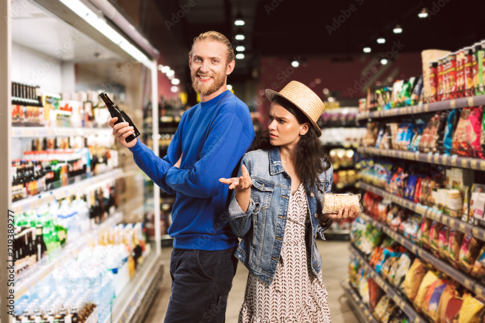Young smiling guy happily looking in camera holding beer in hand while girl thoughtfully looking at him in supermarket