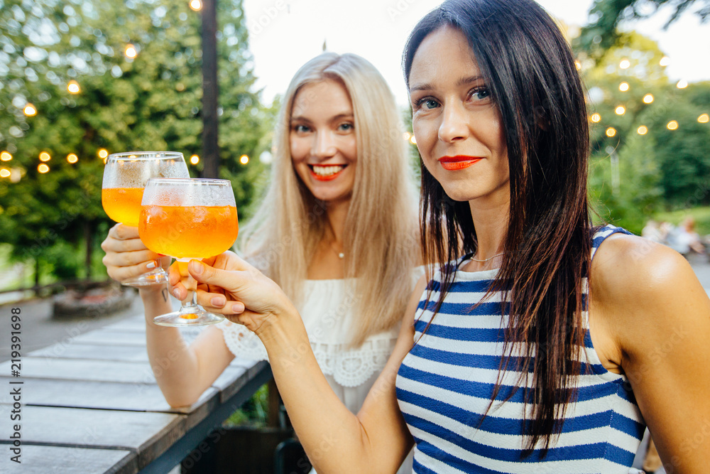 Blond and brunette sweden women drinking aperol spritz, with friend at dinner table in modern outdoor cafe on the green park. Wearing in striped and white dress. Copy space Blond and brunette sweden women drinking aperol spritz, with friend at dinner table in modern outdoor cafe on the green park. Wearing in striped and white dress. Copy space