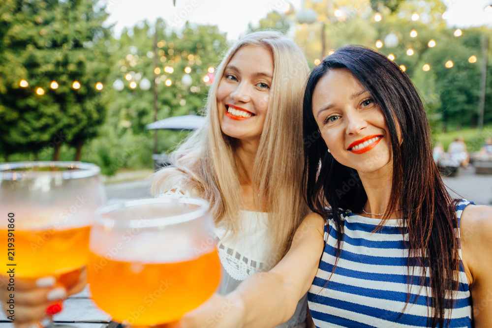 Blond and brunette sweden women drinking aperol spritz, clink glasses with friends, looking at camera in modern outdoor cafe on the green park. Wearing in striped and white dress. Copy space Blond and brunette sweden women drinking aperol spritz, clink glasses with friends, looking at camera in modern outdoor cafe on the green park. Wearing in striped and white dress. Copy space