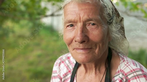 Portrait of an elderly woman in a kerchief. The wind wavers her gray hair, she looks into the camera and blinks. Grandmother in the garden near the house. Close-up