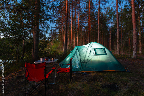 Night landscape with a tent in the forest near lake. The light from the lantern in a tent. Portable table and chairs, green tourist tent. Romantic evening with a tent at night. © korchemkin