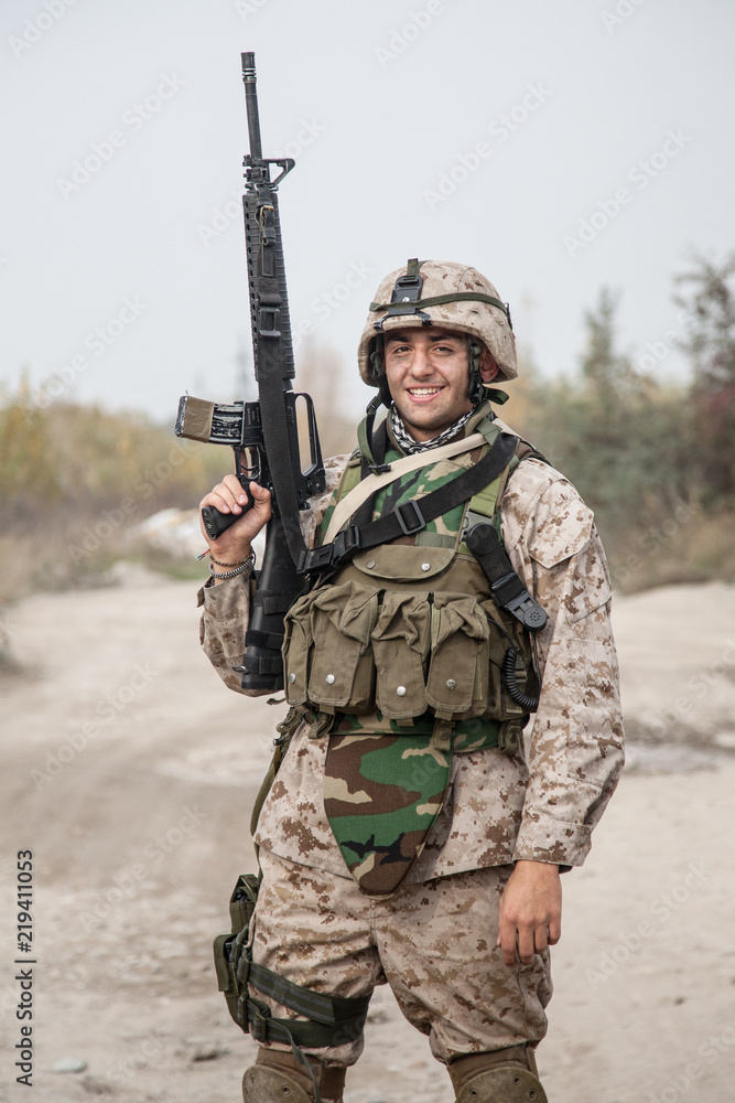 Smiling army soldier, United States Marine Corps infantry shooter in camo  battle uniform, protected with body armor and helmet, posing with assault  rifle in hands while standing near country road foto de