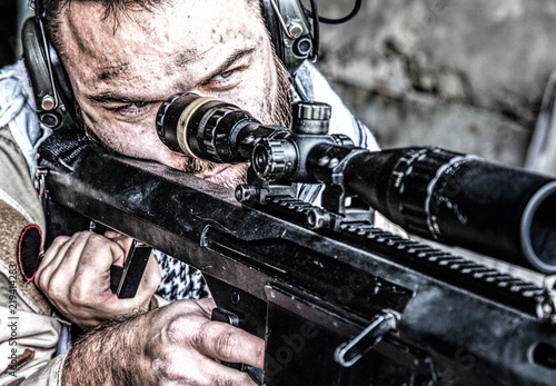 Portrait of U.S. Navy SEAL sniper on firing position, armed with large caliber sniper rifle with telescopic sight, wearing tactical headset with microphone, observing territory, searching targets photo