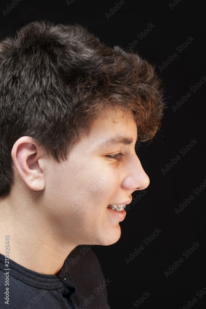 Obraz premium Vertical photo of a guy on a black background in profile. A boy smiles - the braces are clearly visible on the teeth
