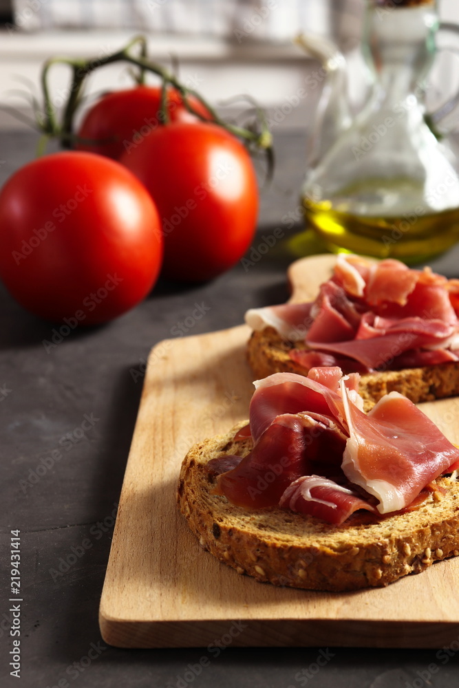 Spanish ham (jamon) on a piece of toasted whole grain bread, tomatoes and olive oil beside