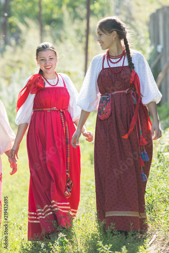 Two young beautiful girls in Russian national dresses are walking on the grass and smiling