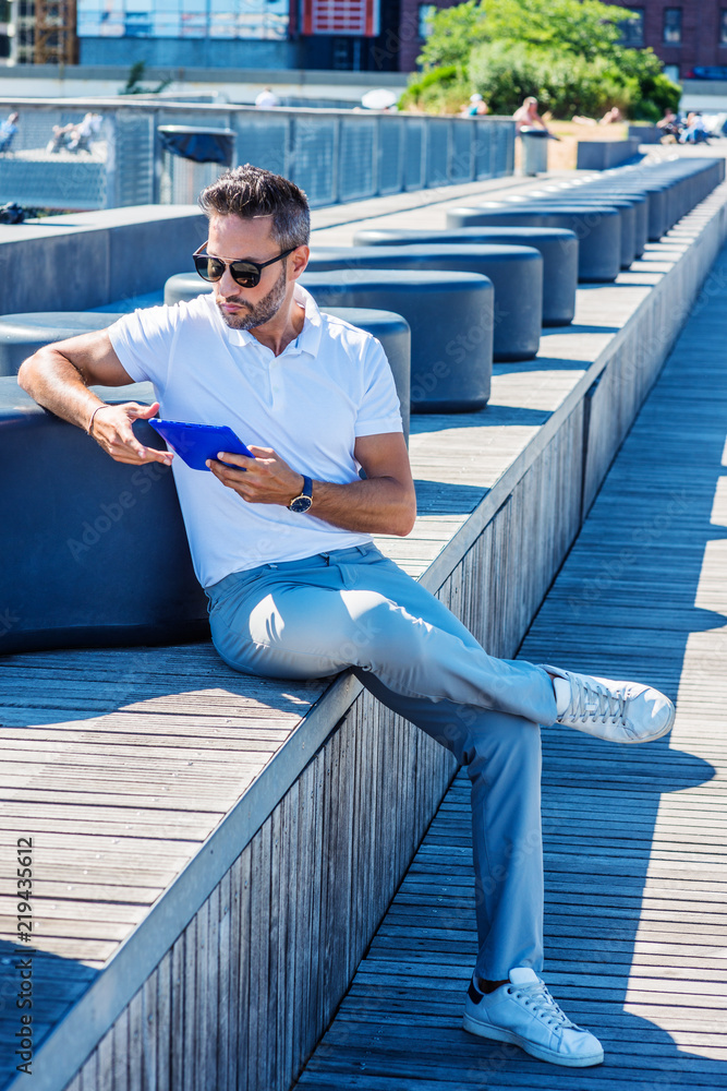 Modern Reading. Young European Man with beard, little gray hair, wearing  white Polo shirt, gray pants, white sneakers, sunglasses, sitting on stairs  at street park in New York, reading tablet computer Stock
