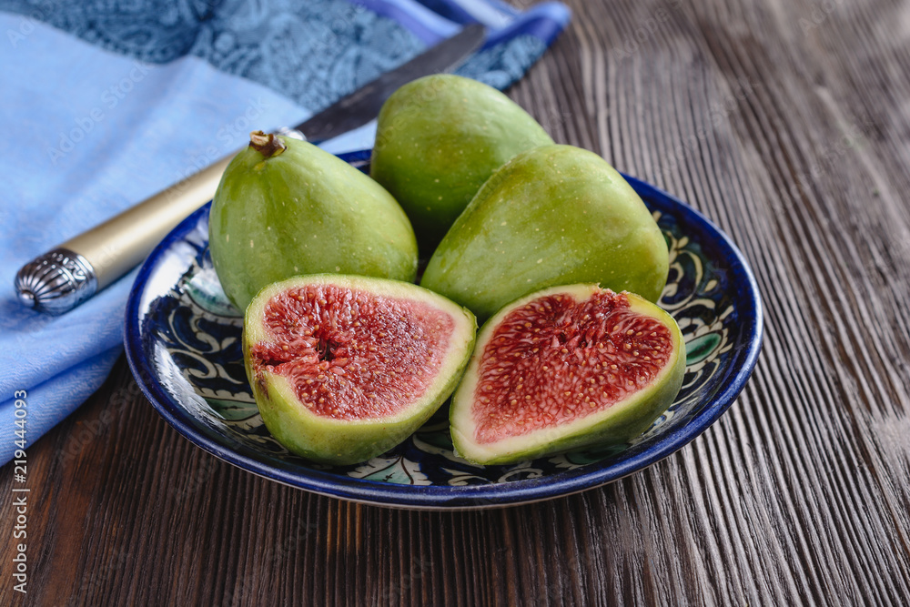 Several ripe figs on a plate with an oriental pattern are on a wooden table next to a blue napkin and a cutting knife, one fig fruit is cut into two halves Several ripe figs on a plate with an oriental pattern are on a wooden table next to a blue napkin and a cutting knife, one fig fruit is cut into two halves