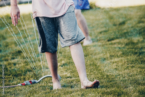 Children playing with the garden sprinkler jumping over with wet clothes. Body parts, close up of legs and feet.